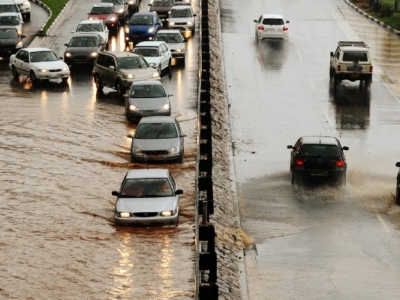 Vehicles driving through muddy floodwater on a divided road, with higher water levels on the left side and partially submerged tires.