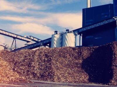 Large piles of wood chips in front of an industrial facility with silos, conveyors, and a smokestack under a partly cloudy sky.