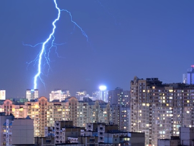 Night cityscape with high-rise buildings illuminated, struck by multiple bright lightning bolts against a dark blue sky.