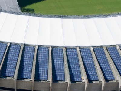Aerial view of a stadium Governador Magalhães Pinto (Mineirão) with rows of solar panels installed on the curved roof, surrounding a green field under a partly cloudy sky.