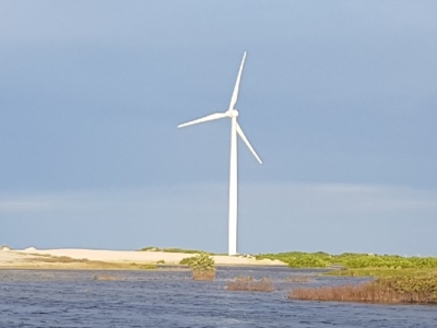 Three large white wind turbines on a sandy coastal landscape with water in the foreground and cloudy sky in the background.
