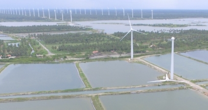 Rows of wind turbines extending across the landscape near rectangular aquaculture ponds, surrounded by greenery and small buildings.