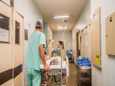 Healthcare worker in scrubs pushing a patient on a hospital stretcher through a brightly lit hallway, with medical equipment and supplies visible along the corridor.