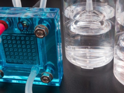 Laboratory setup with a blue electrochemical device connected to red and clear tubing, alongside graduated cylinders partially filled with liquid.