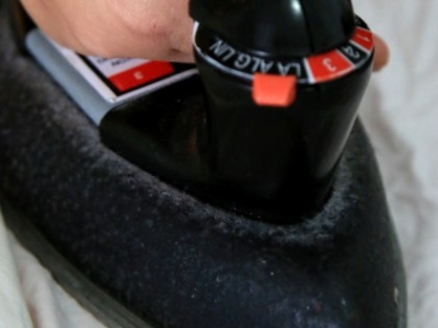 Close-up of a hand gripping a black iron with visible temperature dial, pressing fabric on a flat surface.