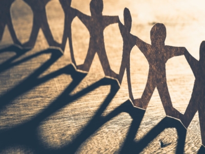 Paper cutout figures holding hands in a chain, casting long shadows on a wooden surface under warm light.