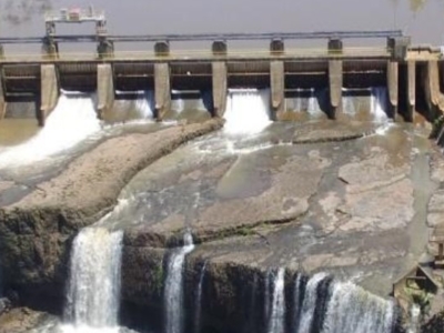 Aerial view of a hydroelectric dam with multiple spillways releasing water, surrounded by green vegetation and access roads.