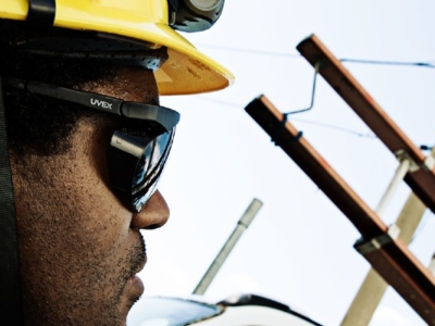Close-up of a worker wearing a yellow safety helmet and protective glasses, observing utility poles and a bucket lift during electrical maintenance.