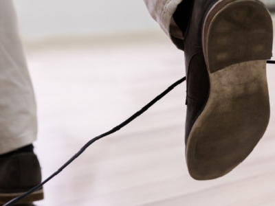 Close-up of a person’s foot stepping over a black electrical cord on a wooden floor, illustrating a common tripping hazard.