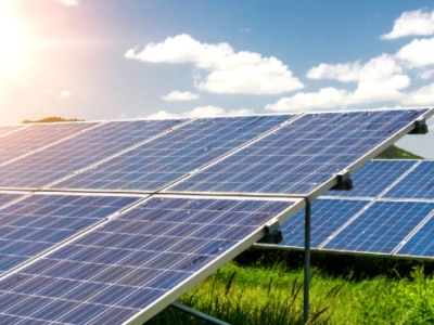 Large outdoor arrays of solar panels on a grassy field under a sunny sky with scattered clouds.