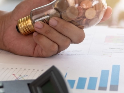 Close-up of a hand holding a light bulb filled with coins, symbolizing financial savings and innovation, with charts and a calculator on a desk.