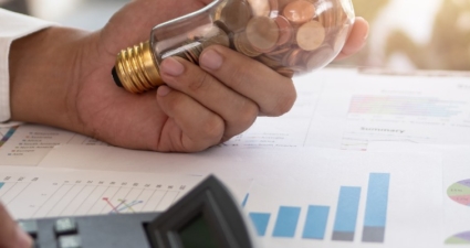 Close-up of a hand holding a light bulb filled with coins, symbolizing financial savings and innovation, with charts and a calculator on a desk.