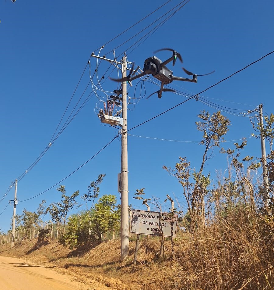 A imagem mostra um drone voando próximo a um poste de energia elétrica em uma estrada de terra.

Cenário: céu azul e limpo, com árvores e vegetação seca nas laterais da estrada.

Elemento principal: o drone está em operação, aparentemente realizando inspeção ou monitoramento da infraestrutura elétrica.

Detalhes adicionais: há um poste com fios de energia e uma placa indicando “ENTRADA E SAÍDA DE VEÍCULOS A 100 M.”.

A cena transmite a ideia de uso de tecnologia em ambiente rural ou semiurbano, voltada para segurança e manutenção de redes elétricas.