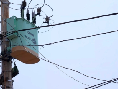 A imagem mostra um transformador elétrico instalado em um poste de utilidade pública. O equipamento é de cor verde clara com a parte inferior em tom bege. No topo, há isoladores de alta tensão conectados a diversos cabos que se estendem horizontal e diagonalmente, distribuindo energia. O texto visível no corpo do transformador diz “CEMIG TRAEL MR”. O fundo é um céu limpo, o que destaca ainda mais os detalhes do transformador e da fiação. Trata-se de parte da infraestrutura de distribuição elétrica que leva energia das linhas principais até residências e estabelecimentos.