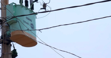 A imagem mostra um transformador elétrico instalado em um poste de utilidade pública. O equipamento é de cor verde clara com a parte inferior em tom bege. No topo, há isoladores de alta tensão conectados a diversos cabos que se estendem horizontal e diagonalmente, distribuindo energia. O texto visível no corpo do transformador diz “CEMIG TRAEL MR”. O fundo é um céu limpo, o que destaca ainda mais os detalhes do transformador e da fiação. Trata-se de parte da infraestrutura de distribuição elétrica que leva energia das linhas principais até residências e estabelecimentos.