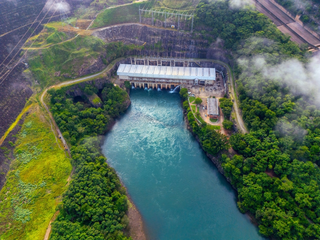 A imagem mostra uma vista aérea de uma usina hidrelétrica em meio a uma paisagem verde e montanhosa. O grande paredão da represa possui comportas por onde a água é liberada para o rio abaixo. Ao lado, há um edifício de telhado metálico que provavelmente abriga as turbinas e geradores, além de construções menores conectadas por estradas internas. Linhas de transmissão partem da instalação, indicando a distribuição da energia produzida. A cena é envolvida por nuvens baixas ou névoa, o que dá profundidade e um aspecto natural ao ambiente, contrastando com a estrutura imponente da engenharia.