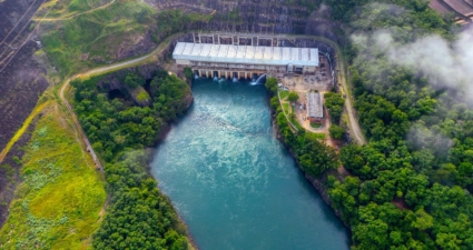 A imagem mostra uma vista aérea de uma usina hidrelétrica em meio a uma paisagem verde e montanhosa. O grande paredão da represa possui comportas por onde a água é liberada para o rio abaixo. Ao lado, há um edifício de telhado metálico que provavelmente abriga as turbinas e geradores, além de construções menores conectadas por estradas internas. Linhas de transmissão partem da instalação, indicando a distribuição da energia produzida. A cena é envolvida por nuvens baixas ou névoa, o que dá profundidade e um aspecto natural ao ambiente, contrastando com a estrutura imponente da engenharia.