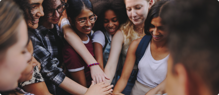 Um grupo de jovens sorrindo, homens e mulheres de diversas etnias, está reunido em círculo, com as mãos empilhadas no centro.
