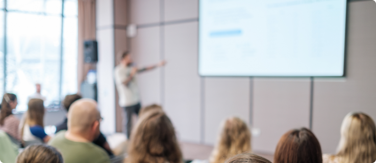 Uma sala de conferências ou sala de aula em foco suave. No fundo, um palestrante está de pé em frente a uma tela de projeção branca, apontando para ela. A audiência, com a cabeça e os ombros visíveis na frente, está sentada em cadeiras assistindo à apresentação. A iluminação é difusa e as paredes têm painéis geométricos.