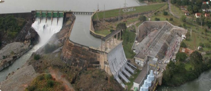 Vista aérea de uma usina hidrelétrica com vertedouros abertos e estrutura da estação ambiental de Itutinga ao lado.