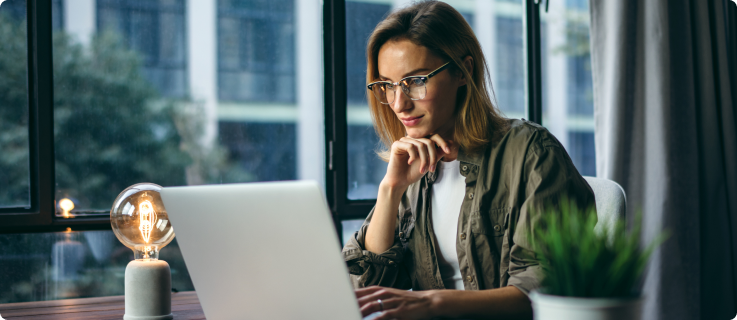 Mulher usando óculos e jaqueta verde-oliva trabalha concentrada em um notebook, sentada em uma mesa com uma luminária acesa e um vaso de planta. Ao fundo, janelas mostram um prédio e árvores do lado de fora.