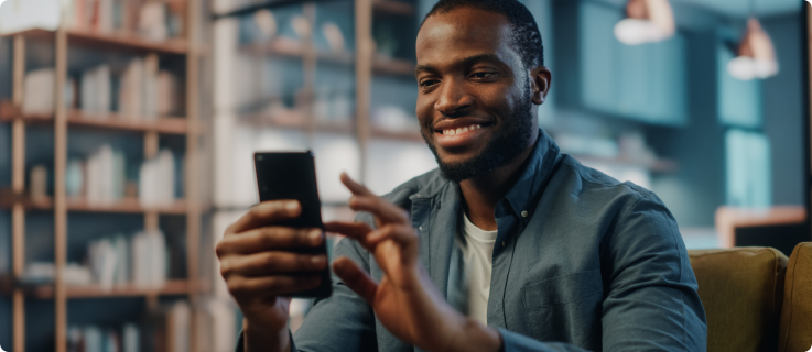 Homem jovem sorridente mexe no celular em um ambiente moderno com estante de livros ao fundo.