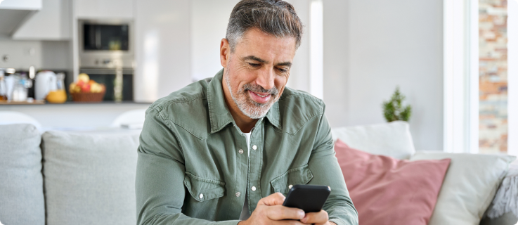 Homem de meia-idade, com barba grisalha e camisa verde, sentado em um sofá enquanto olha para o celular, em uma sala clara e aconchegante.