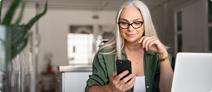 Mulher de cabelos grisalhos e óculos escuros sorri levemente enquanto olha para um celular. Ela está sentada à mesa, com um notebook aberto à frente, em um ambiente claro e moderno.