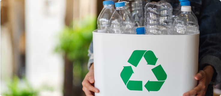 Pessoa segurando uma lixeira branca com símbolo de reciclagem verde, contendo garrafas plásticas.