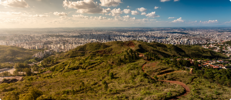 Uma paisagem panorâmica de Belo Horizonte vista de uma colina com vegetação rasteira em primeiro plano. A cidade se estende por um vasto vale, com arranha-céus no centro e áreas residenciais mais densas nas extremidades, sob um céu parcialmente nublado ao entardecer.