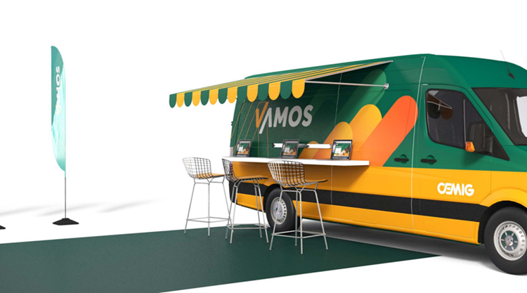 Unidade móvel da Cemig com identidade visual verde, amarela e laranja, com a palavra “VAMOS” na lateral. A van tem toldo, balcão com notebooks e cadeiras altas, simulando um atendimento externo. À esquerda, há uma bandeira vertical com o mesmo tema visual.