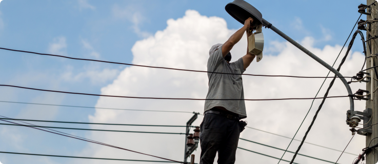 Trabalhador em altura realizando a manutenção de um poste de iluminação pública, com fios elétricos ao fundo e céu parcialmente nublado.