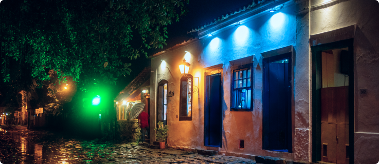 Rua de paralelepípedo com casas coloniais iluminadas por luzes coloridas e postes à noite.
