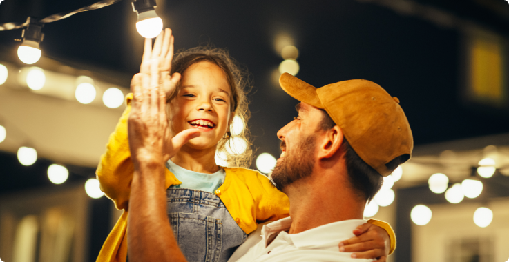 Pai e filha sorrindo e batendo palmas sob cordões de luzes decorativas, em um ambiente externo à noite.