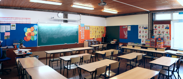 Sala de aula com carteiras organizadas, quadro verde e decoração colorida nas paredes, iluminada por lâmpadas fluorescentes.