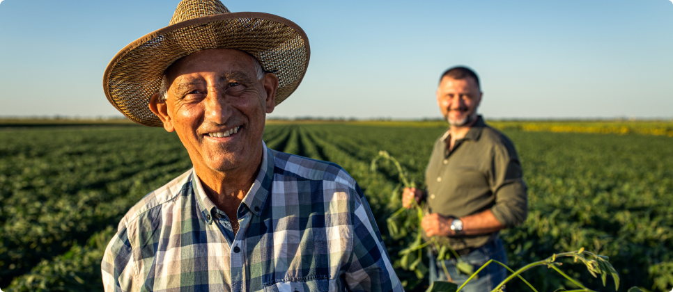 Dois agricultores em uma plantação ao ar livre durante o dia. Um homem mais velho, sorridente e de chapéu de palha, está em destaque na frente. Ao fundo, outro homem segura uma planta e sorri. Ambos estão cercados por fileiras de cultivo verde sob um céu claro.