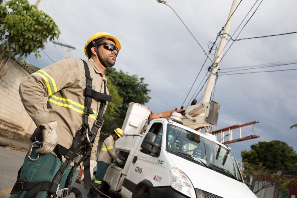 A imagem mostra trabalhadores de utilidade pública realizando manutenção elétrica em uma rua. Eles estão usando equipamentos de segurança — capacetes, luvas e cintos de proteção — próximos a um caminhão equipado com um braço mecânico (boom lift).

Um dos trabalhadores aparece em primeiro plano olhando para cima, provavelmente avaliando os cabos elétricos, enquanto outro está próximo ao veículo. O caminhão traz inscrições como “7340” e “MINAS GERAIS”, indicando que pertence a uma companhia de energia da região.

Ao fundo, vemos postes e fiação elétrica, reforçando o contexto de trabalho em infraestrutura urbana. A cena transmite a importância das medidas de segurança e da preparação cuidadosa em ambientes de risco.