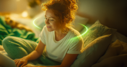Uma jovem ruiva sorrindo enquanto usa um notebook na cama, com um anel de luz verde neon brilhante circulando ao redor de seus ombros em um quarto com iluminação quente e aconchegante.
