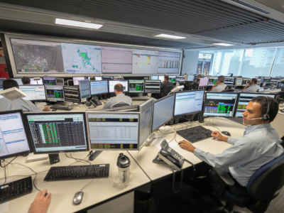 A imagem mostra uma sala de controle moderna, organizada em fileiras de estações de trabalho. Cada estação possui vários monitores exibindo dados, gráficos, mapas e tabelas coloridas. Na parede frontal, há grandes telas que apresentam mapas com informações geográficas ou meteorológicas, além de gráficos e quadros de status. Algumas pessoas estão sentadas nas mesas, trabalhando nos computadores e utilizando telefones ou headsets, o que sugere um ambiente de monitoramento em tempo real. O espaço transmite a ideia de coordenação complexa e análise contínua de informações, típico de operações ligadas a logística, redes ou segurança.