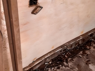 A imagem mostra uma parede interna bastante danificada, provavelmente por fogo ou água. Há um buraco no qual ficam visíveis fios elétricos expostos e uma tampa de tomada solta, pendurada. A parte inferior da parede e o piso estão manchados e cobertos por detritos, sugerindo que houve um evento destrutivo significativo. O ambiente transmite a ideia de um espaço em reparo ou inspeção, possivelmente após um acidente ou desastre, com foco em avaliar danos estruturais e elétricos.