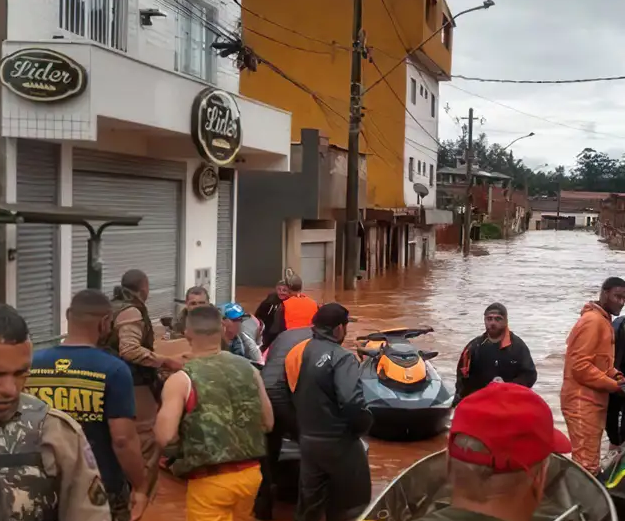 A mensagem transmitida pela imagem é de alerta e solidariedade diante de uma situação de desastre natural.

Elementos principais da mensagem
Impacto da enchente: As ruas estão completamente alagadas, mostrando a gravidade da inundação e como ela afeta diretamente a vida urbana.

Resposta emergencial: A presença de equipes de resgate e equipamentos como o jet ski reforça a ideia de mobilização imediata para salvar vidas e oferecer apoio.

Comunidade vulnerável: Pessoas reunidas em frente ao comércio fechado (“Líder”) evidenciam como o cotidiano foi interrompido e como moradores dependem da ajuda externa.

Urgência e solidariedade: A cena transmite a necessidade de união, preparação e resposta rápida em momentos críticos.