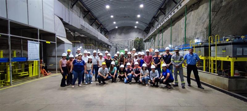Grupo de homens e mulheres posa para foto dentro da Usina Hidrelétrica UHE Queimado, localizada em Unaí MG. A maioria das pessoas usa capacetes de segurança brancos, algumas vestem coletes ou uniformes de trabalho. O grupo está organizado em pé e agachado no centro do espaço. Ao redor, há máquinas industriais, tubulações amarelas e azuis, painéis metálicos e paredes de rocha exposta, sob um teto curvo com iluminação artificial.