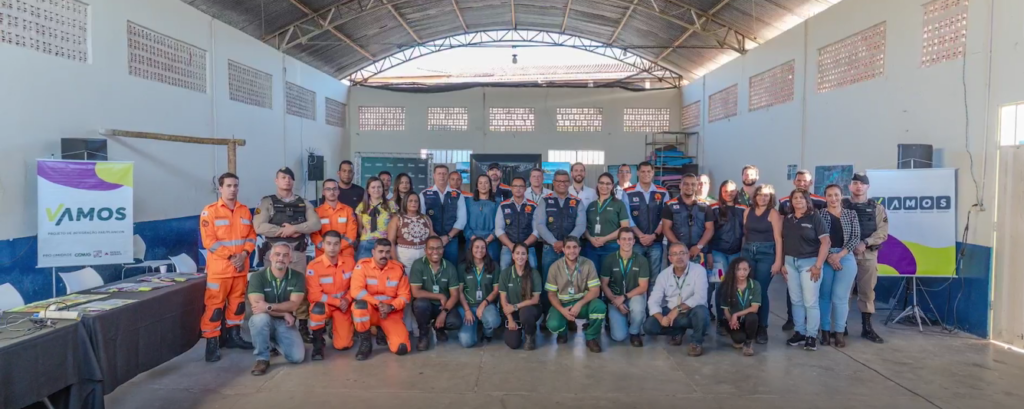 Grupo grande de pessoas posa para foto em um galpão coberto, com paredes claras e estrutura metálica no teto. Há homens e mulheres de diferentes idades, organizados em pé e agachados, alguns usando uniformes laranja de resgate, outros coletes, camisetas verdes e roupas civis. À esquerda e à direita aparecem banners com a palavra “VAMOS”.