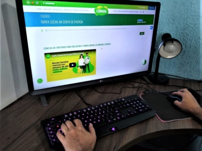 Monitor de computador sobre uma mesa exibindo a página da Cemig sobre Tarifa Social na conta de energia. Em primeiro plano, uma pessoa digita em um teclado com iluminação roxa e usa o mouse ao lado, indicando navegação no site. Ao lado do monitor há um abajur preto.