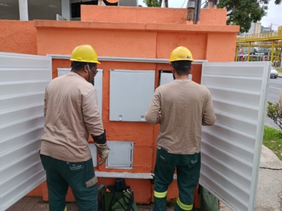 A imagem mostra dois trabalhadores de utilidade pública realizando manutenção em um painel elétrico ou de serviços. Eles estão usando capacetes de segurança amarelos e roupas de proteção. O painel, instalado em uma parede laranja, tem portas brancas abertas que revelam vários compartimentos internos. Um dos trabalhadores está manuseando ferramentas retiradas de uma bolsa verde colocada no chão, enquanto o outro parece inspecionar ou ajustar componentes dentro do painel. O cenário é externo, próximo a uma calçada e a uma rua, com infraestrutura urbana visível ao fundo, incluindo prédios e uma passarela para pedestres.