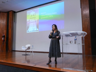 Mulher falando em um palco com microfone, com a tela de projeção ao fundo exibindo a capa do livro "Um Poema para Bárbara" e o título "Minha Vida/Poema para Bárbara/TRF6".