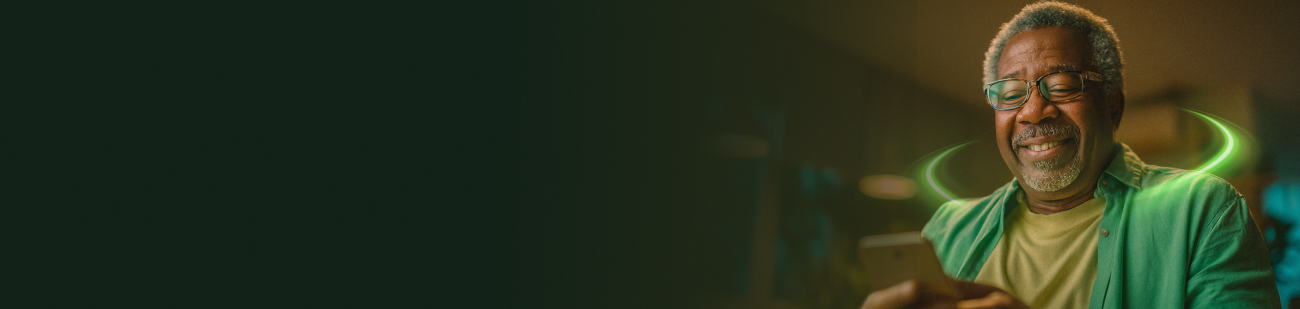 Um homem negro de meia-idade, com cabelos e barba grisalhos, sorri enquanto olha para a tela de um smartphone que segura com as duas mãos. Ele usa óculos de grau e uma camisa verde sobre uma camiseta amarela. Ao redor de seus ombros, há um efeito visual de anéis de luz neon verde brilhante em movimento. O fundo é um ambiente interno aconchegante e levemente desfocado, com iluminação quente.