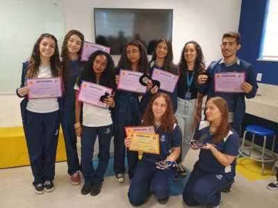 imagem mostra um grupo de nove pessoas reunidas em um ambiente interno, provavelmente uma sala de aula ou espaço educacional. Oito delas parecem ser estudantes, vestindo uniformes escolares, e uma pessoa — possivelmente instrutor ou coordenador — usa um crachá. Os estudantes seguram certificados coloridos, em sua maioria roxos e um amarelo, com a palavra “CERTIFICADO” impressa no topo, além de pequenos prêmios circulares. O cenário inclui um quadro branco, uma televisão e um balcão com banquetas ao fundo, reforçando o clima acadêmico. A cena transmite claramente um momento de reconhecimento e celebração de conquistas estudantis, com os alunos sendo premiados por seu desempenho.