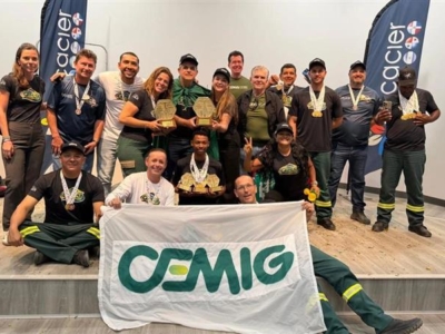 A imagem mostra um grupo de pessoas reunidas em um palco, todas vestindo uniformes e exibindo medalhas. Alguns seguram troféus, o que indica que participaram de uma competição ou cerimônia de premiação. Na frente, duas pessoas seguram uma faixa branca com o logotipo verde e amarelo da CEMIG. Ao fundo, há dois banners verticais com a palavra Cacier e outros logotipos. O ambiente é claramente de um evento formal, voltado para reconhecimento e celebração de conquistas coletivas.