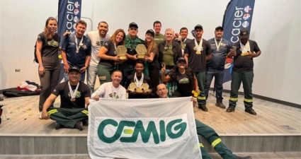A imagem mostra um grupo de pessoas reunidas em um palco, todas vestindo uniformes e exibindo medalhas. Alguns seguram troféus, o que indica que participaram de uma competição ou cerimônia de premiação. Na frente, duas pessoas seguram uma faixa branca com o logotipo verde e amarelo da CEMIG. Ao fundo, há dois banners verticais com a palavra Cacier e outros logotipos. O ambiente é claramente de um evento formal, voltado para reconhecimento e celebração de conquistas coletivas.